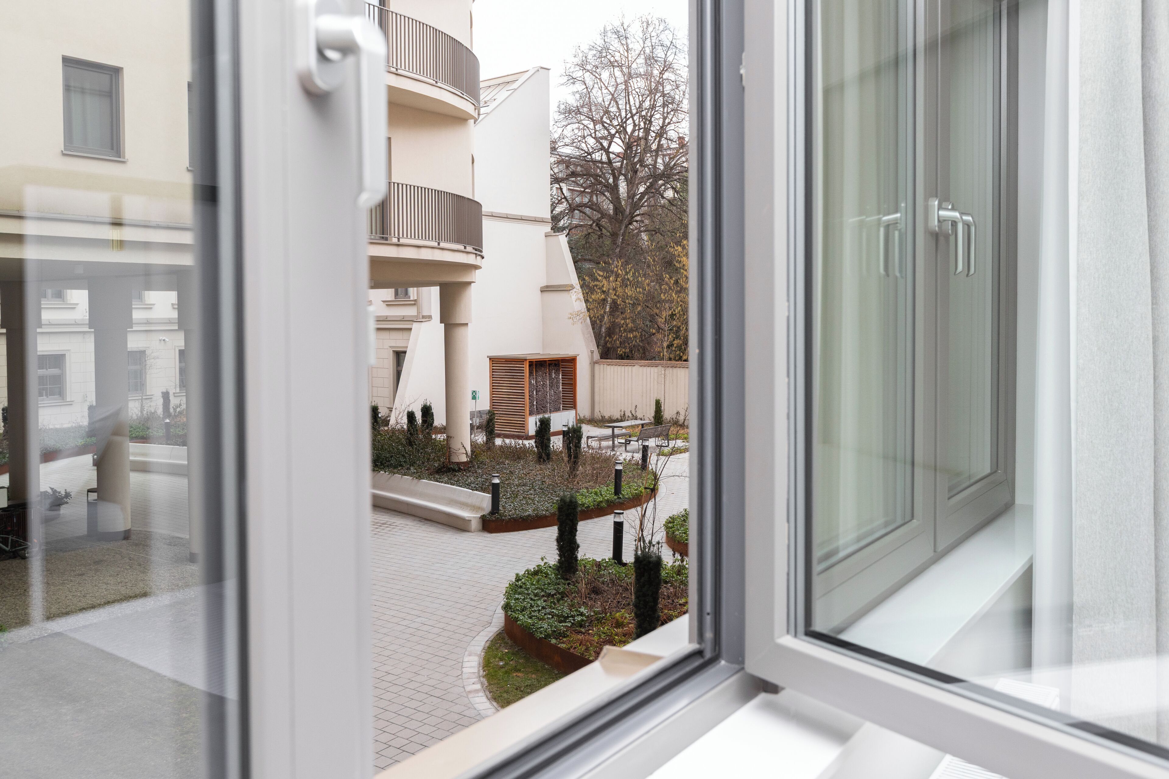 Vista a través de una ventana que muestra un patio con un edificio, un árbol y un banco.