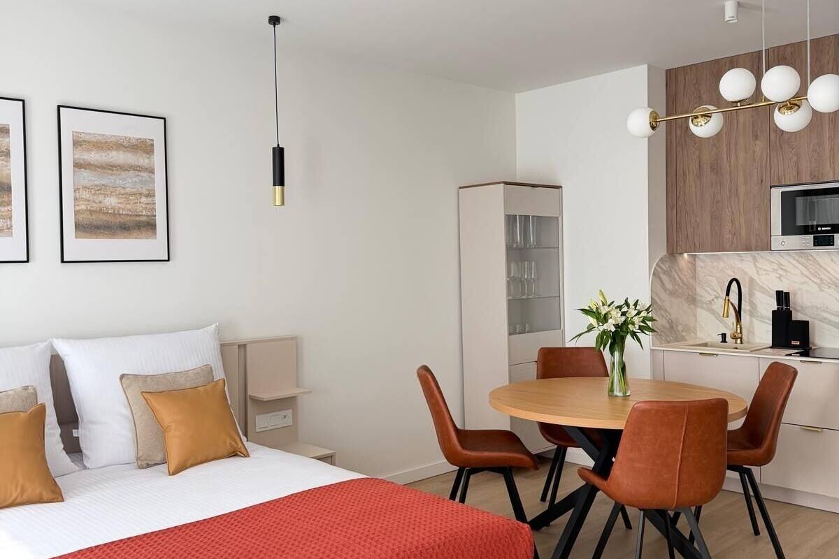 A modern hotel room with a bed, a dining table with chairs, a kitchen area, and a TV.