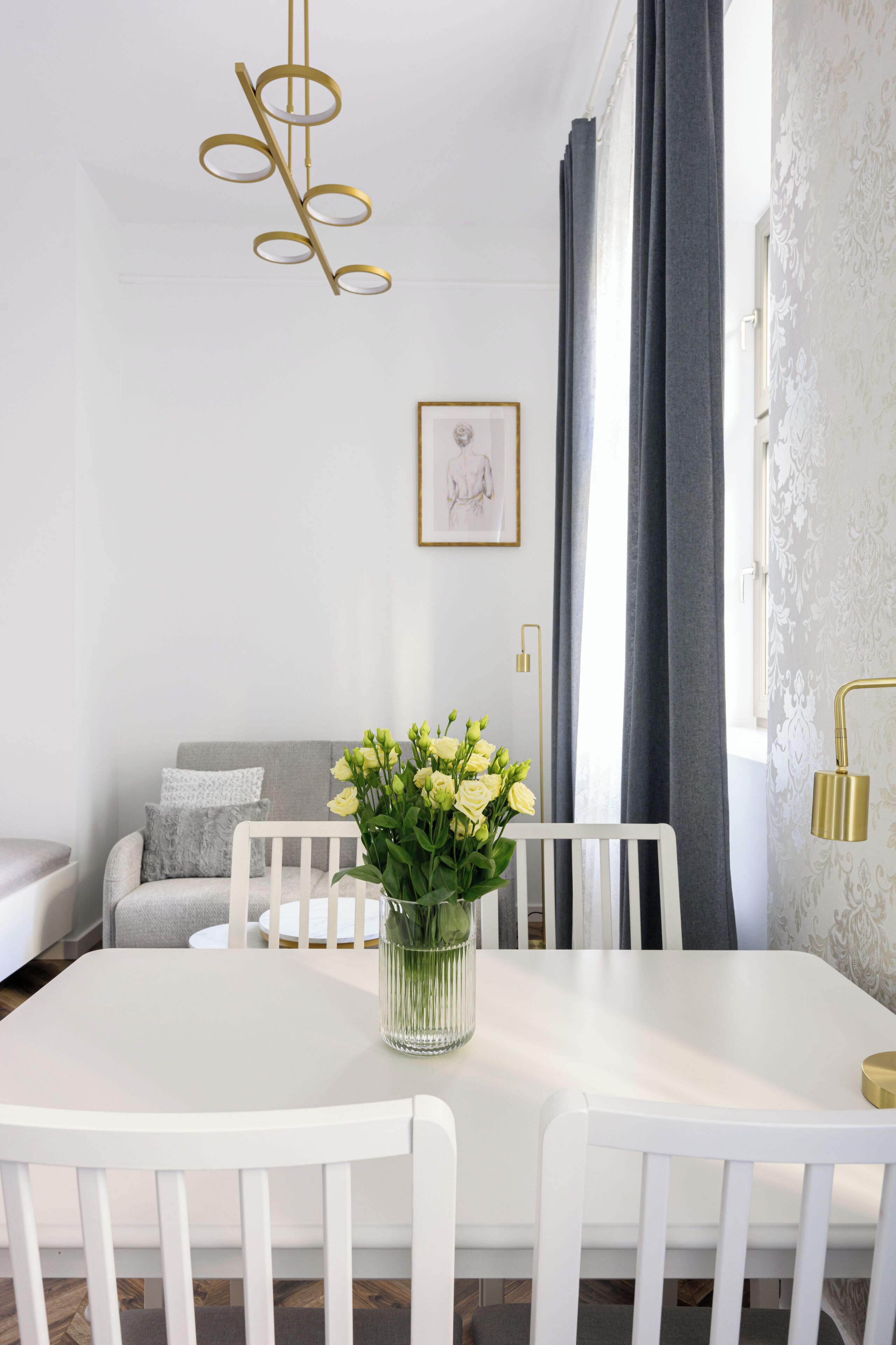 A dining area with a white table, chairs, and a vase with flowers. There is a sofa with pillows and a framed picture on the wall.