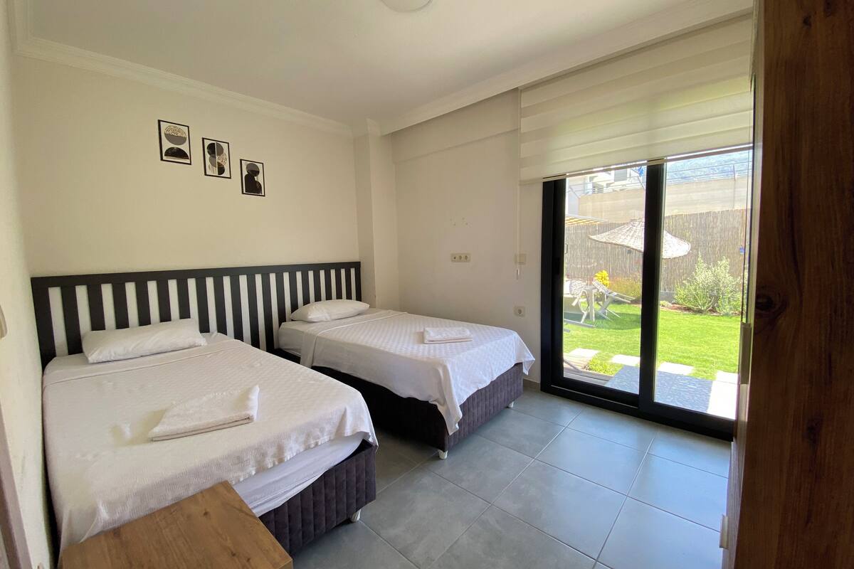 Une chambre avec deux lits, une table de chevet en bois et une porte-fenêtre coulissante donnant sur un jardin.
