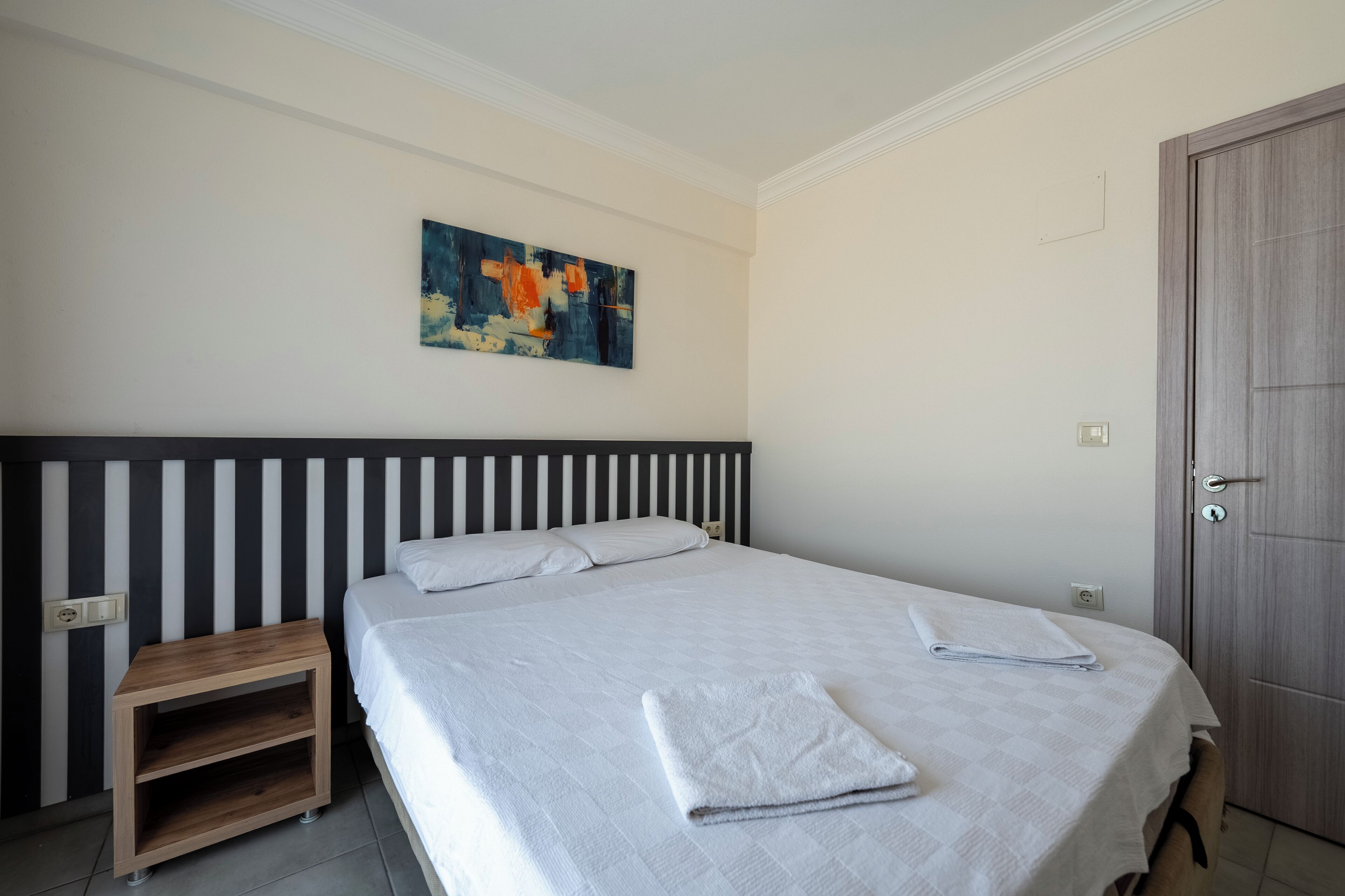 A neatly made bed with a white bedspread and a folded towel, a wooden nightstand with a drawer, a wall-mounted abstract painting, and a wooden door.