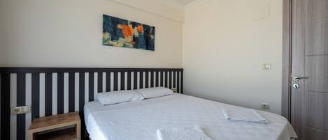 A neatly made bed with a white bedspread and a folded towel, a wooden nightstand with a drawer, a wall-mounted abstract painting, and a wooden door.