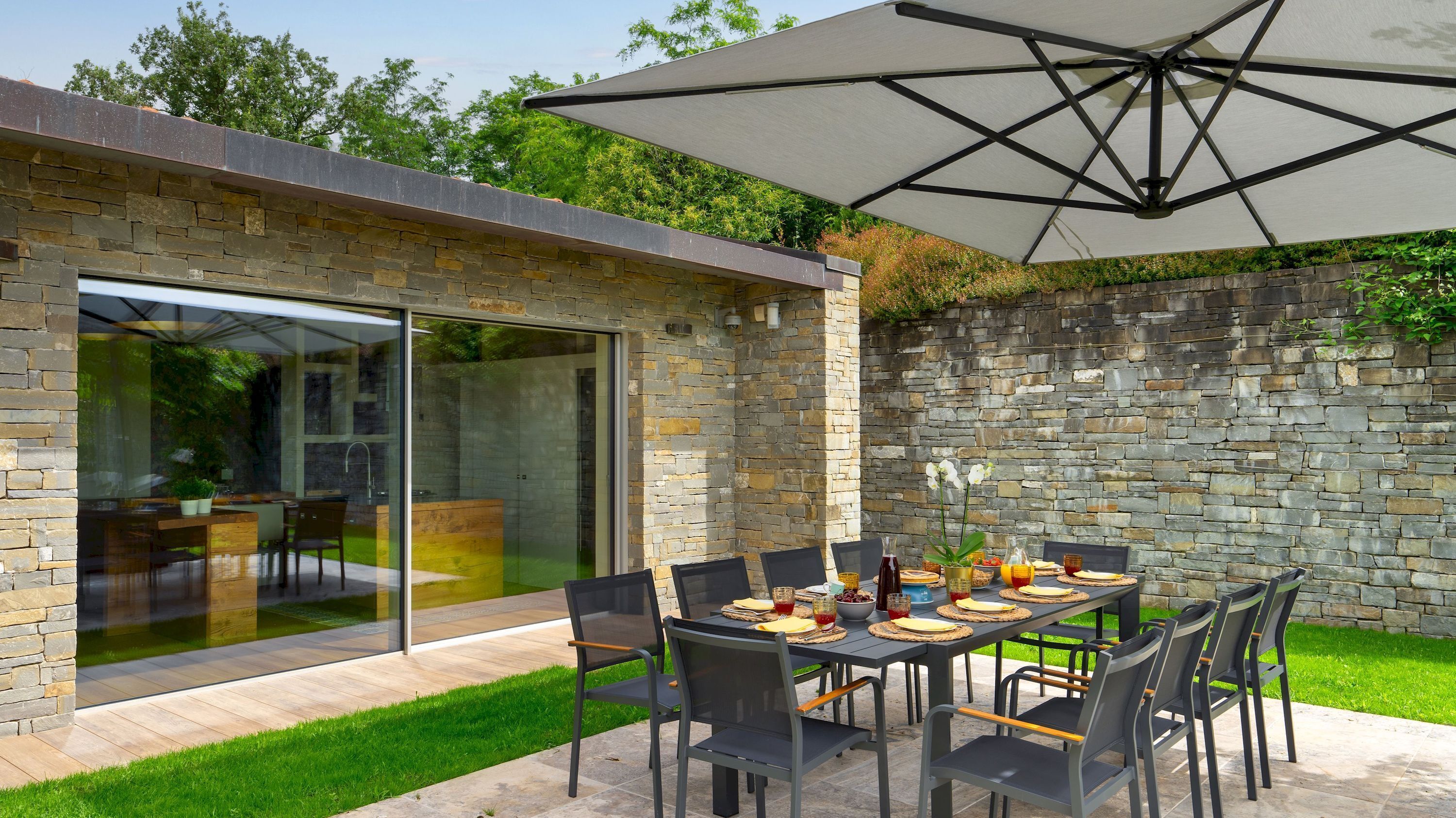 An outdoor dining area with a stone wall, a glass door leading to a patio, and a large umbrella providing shade.