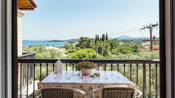 A balcony with a table set for two, overlooking a coastal landscape with trees and buildings.