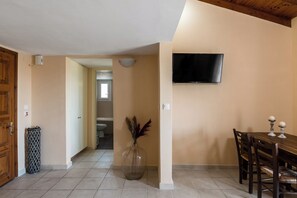 A room with a wooden door, a trash bin, a vase with dried plants, a dining table with chairs, and a TV mounted on the wall.