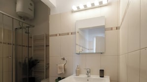 A bathroom with a glass shower enclosure, a white sink, a mirror with lights, and a towel rack.