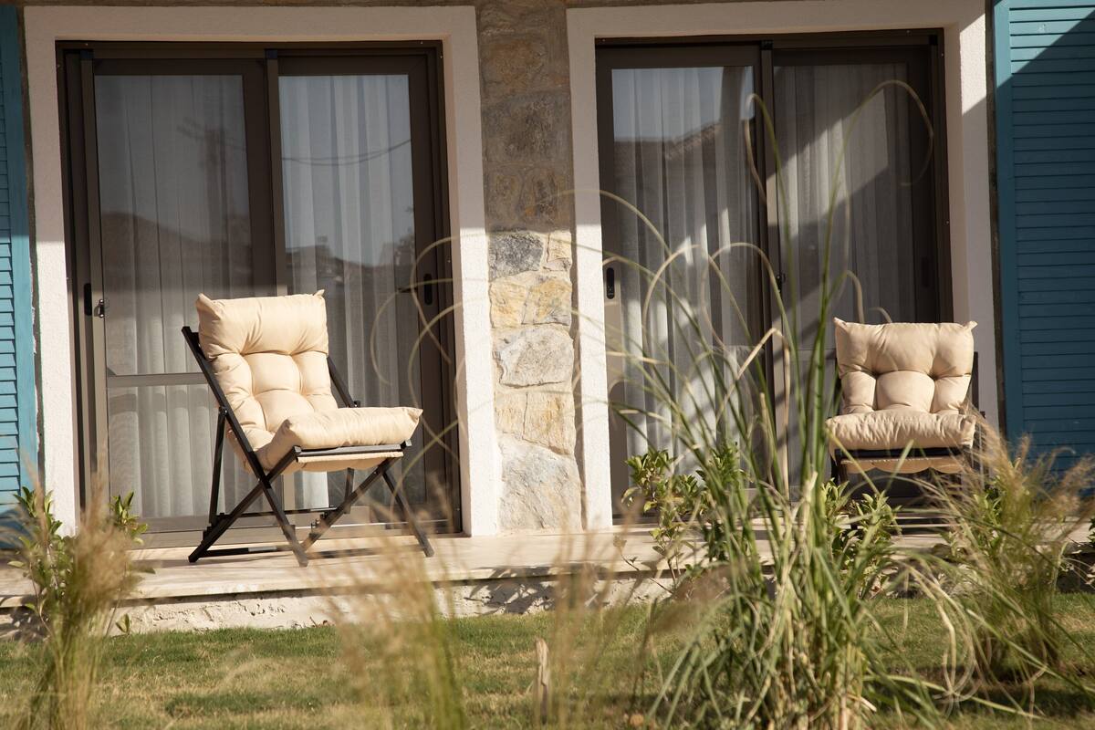 Two cushioned chairs on a porch with stone pillars and a view of a house with blue siding.