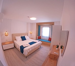 A modern hotel room with a large bed, bedside tables, a small seating area with cushions, a window with curtains, and a mirror. - Eski Kapı Datça  (Datça)