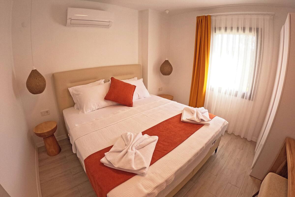 A neatly made bed with a red blanket, white pillows, and a folded towel. There is a wooden nightstand with a lamp, a wooden chair, and a window with curtains.