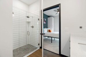 A modern bathroom with a glass shower enclosure, a white sink, and a door leading to a bedroom with a bed and a ceiling fan.