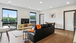 Una sala de estar moderna con un sofá negro, suelo de madera, un televisor y ventanas amplias que dan a zonas verdes.