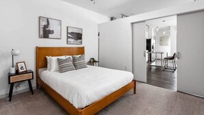 Un dormitorio con cama de madera, ropa de cama blanca, dos cuadros en la pared, un ventilador de techo y vistas al comedor contiguo.