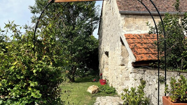 A rustic stone house with a tiled roof, a wooden archway with 'COSY OURCQ' written on it, a stone staircase leading to a door, and a garden with greenery and a potted plant. - Cosy sur Ourcq (TROESNES)