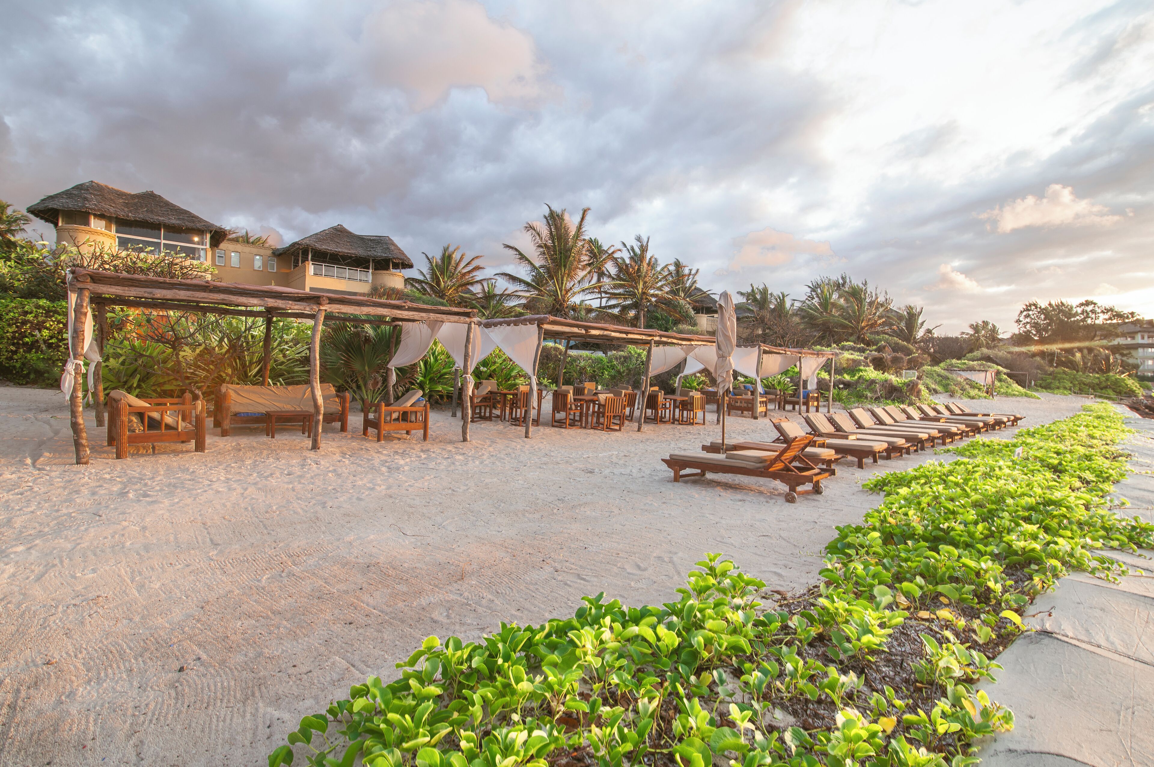 Un complexe hôtelier en bord de mer, doté de chaises longues et de tables en bois, abritées sous des toits de chaume, et entouré d’une végétation luxuriante et de palmiers.