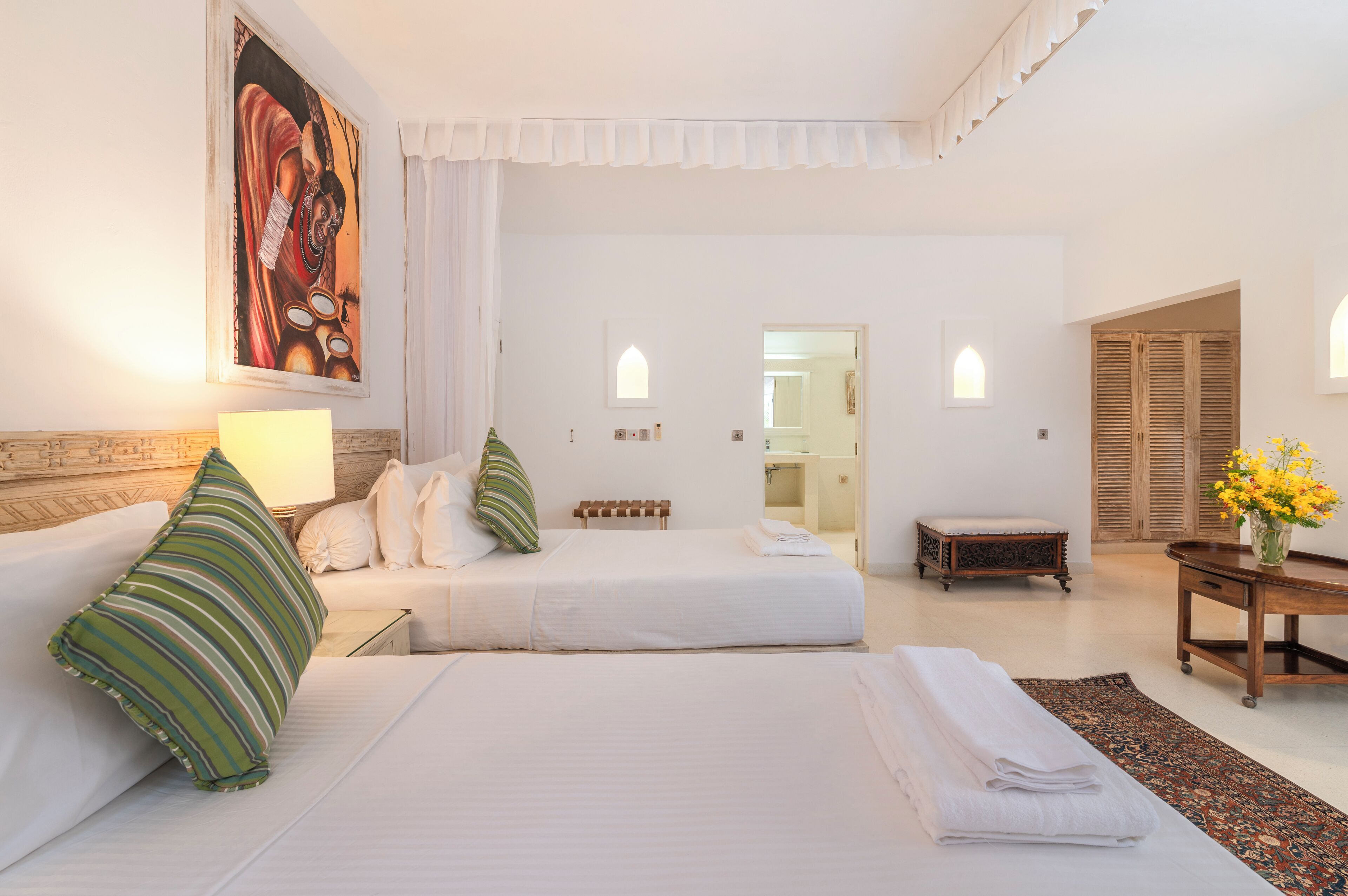 A hotel room with a bed, a bench, a table with a vase of flowers, and a painting on the wall.