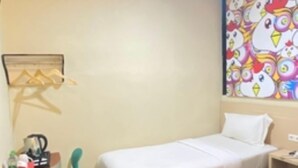 Une chambre d’hôtel avec deux lits, un bureau avec une chaise, une petite table avec une bouilloire et une décoration murale colorée sur le thème des poulets.