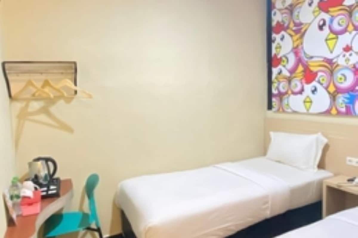 Ein Hotelzimmer mit zwei Betten, einem Schreibtisch mit Stuhl, einem kleinen Tisch mit Wasserkocher und einer farbenfrohen Wanddekoration mit Hühnern.