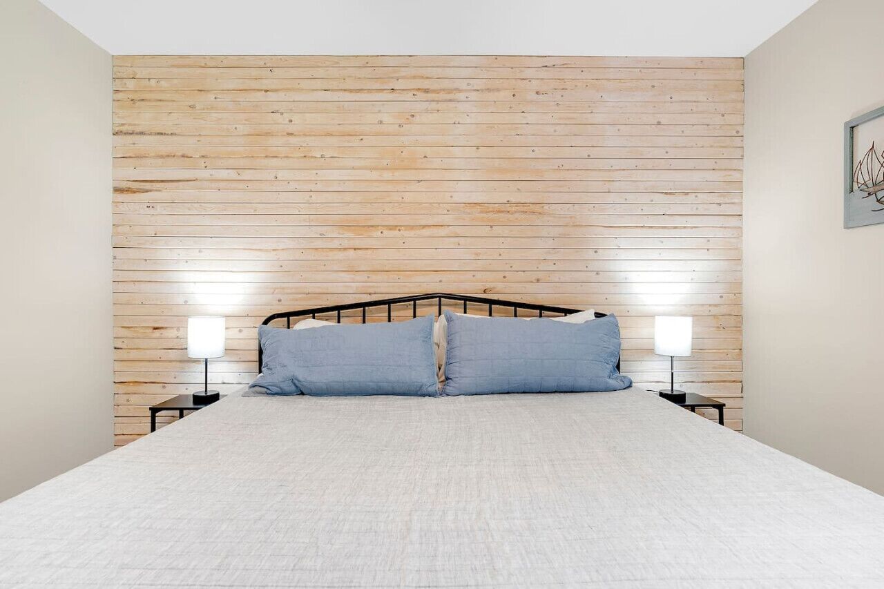 A bedroom with a wooden slat headboard, a bed with white bedding, two bedside tables with lamps, and a framed picture on the wall.