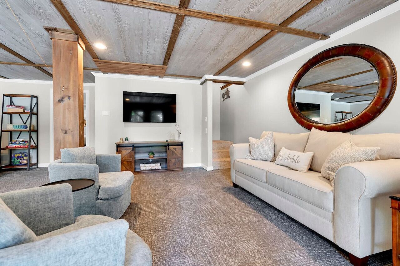 A living room with a white sofa, grey armchairs, a round mirror, and a flat-screen TV mounted on the wall.