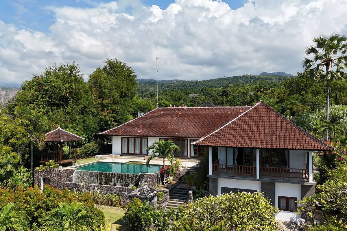 Une maison à deux étages avec une piscine, entourée d’une végétation luxuriante et adossée à un paysage montagneux.