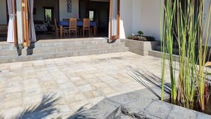 A covered patio area with stone flooring, wooden steps, and a dining area visible through open curtains. - Ponjok Batu Residence (Tejakula)