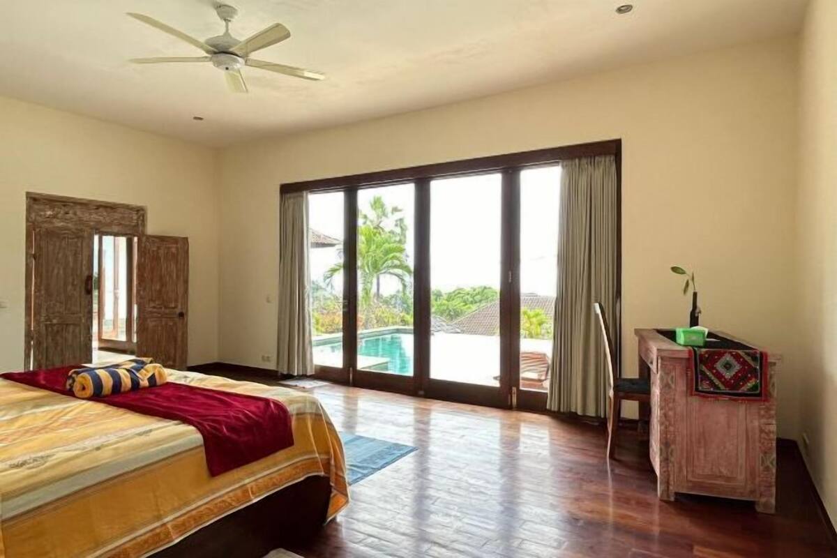Une chambre d’hôtel équipée d’un lit, d’un bureau, d’une chaise, d’un ventilateur de plafond et d’une grande porte-fenêtre coulissante donnant sur un espace piscine.