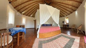 A bedroom with a thatched roof, a bed with a canopy, a dining table with chairs, and a small table with a lamp. - Ponjok Batu Residence (Tejakula)