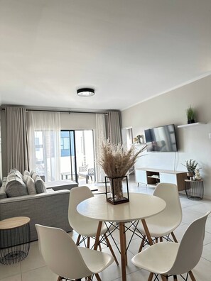 Uma sala de estar moderna com uma mesa de jantar redonda, cadeiras brancas, um sofá e uma TV na parede.