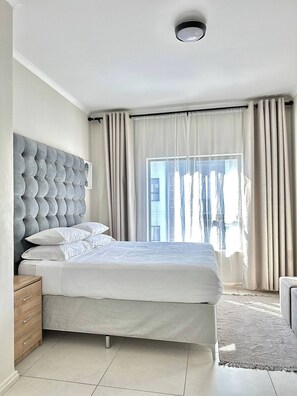 Relax in a serene bedroom with a large bed and tufted headboard, surrounded by sheer curtains and a tiled floor.