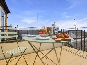 Een ronde tafel met borden, een schaal aardbeien en croissants op een balkon met uitzicht op een steiger en water.