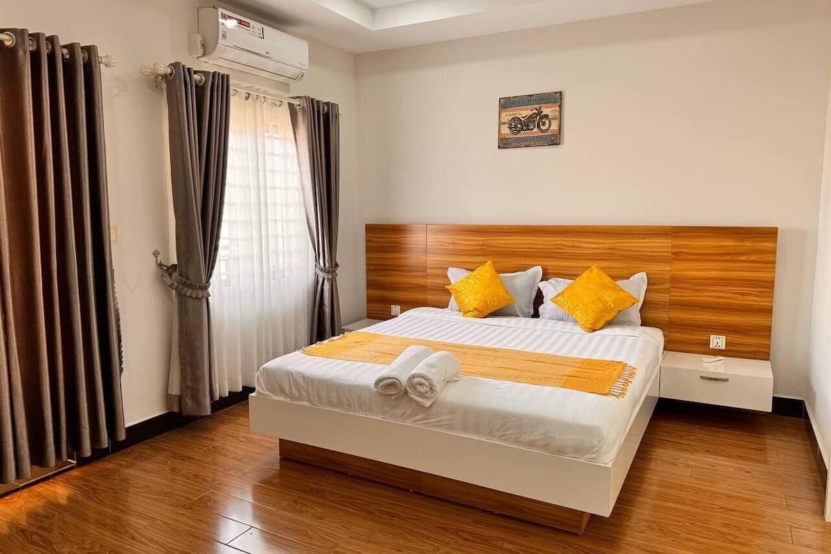 A hotel room with a wooden headboard, a bed with white bedding and yellow accents, a window with curtains, and an air conditioning unit.