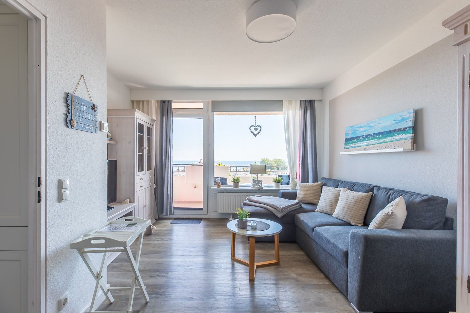 A cozy living room with a grey sofa, a small round table, and a view of the sea through a balcony door.