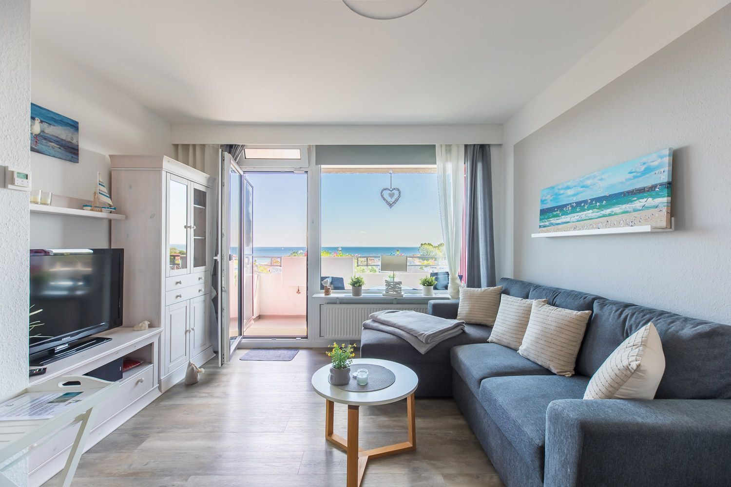 A modern living room with a grey sofa, a flat-screen TV, and a balcony with a view of the sea.
