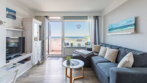 Una sala de estar moderna con un sofá gris, un televisor de pantalla plana y un balcón con vistas al mar.
