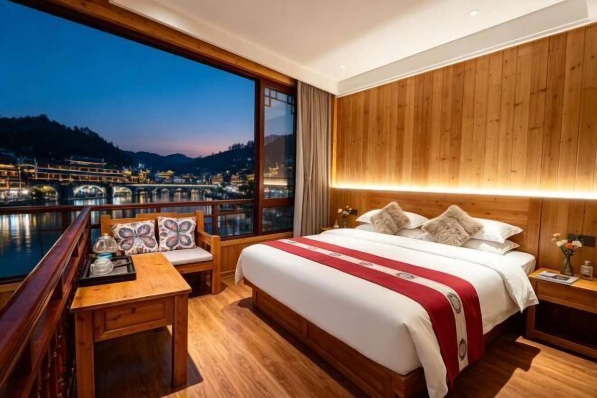 Ein Hotelzimmer mit großem Fenster, einem Bett mit rotem und weißem Überwurf, Holzvertäfelung an den Wänden und Blick auf einen Fluss und Gebäude im Dämmerlicht.