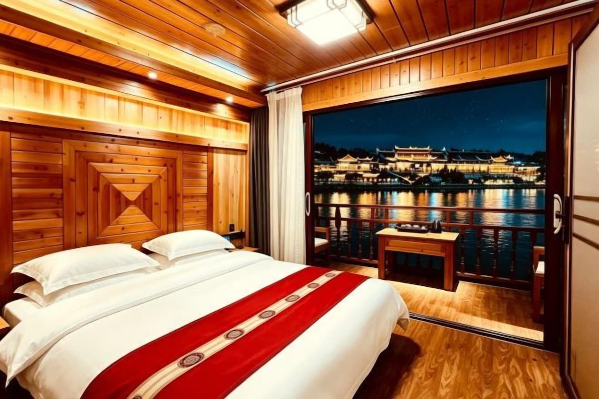 A wooden-paneled room with a bed, a small table, and a view of a water body with buildings at night.