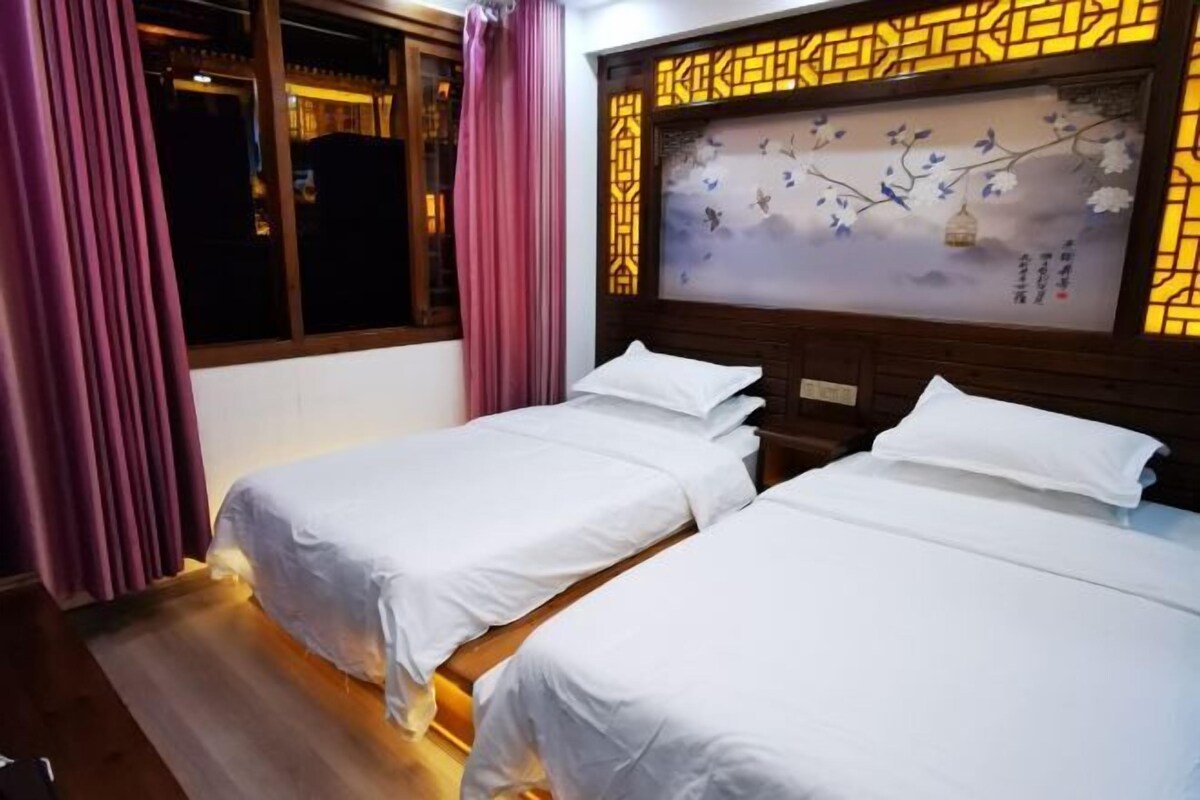 A hotel room with two single beds, wooden flooring, a window with curtains, and a decorative wall panel.