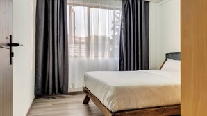 A bedroom with a wooden bed, dark curtains, and a view of buildings through sheer curtains. - Affordable Spectacular Urban Stays (Nairobi)