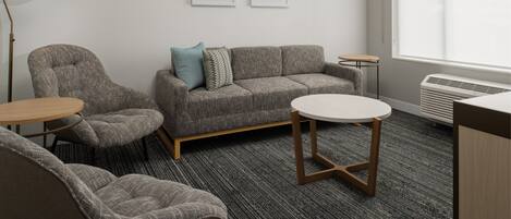 A modern room with a grey sofa, two armchairs, a round table, and a standing lamp. There are two framed artworks on the wall and a window with blinds.