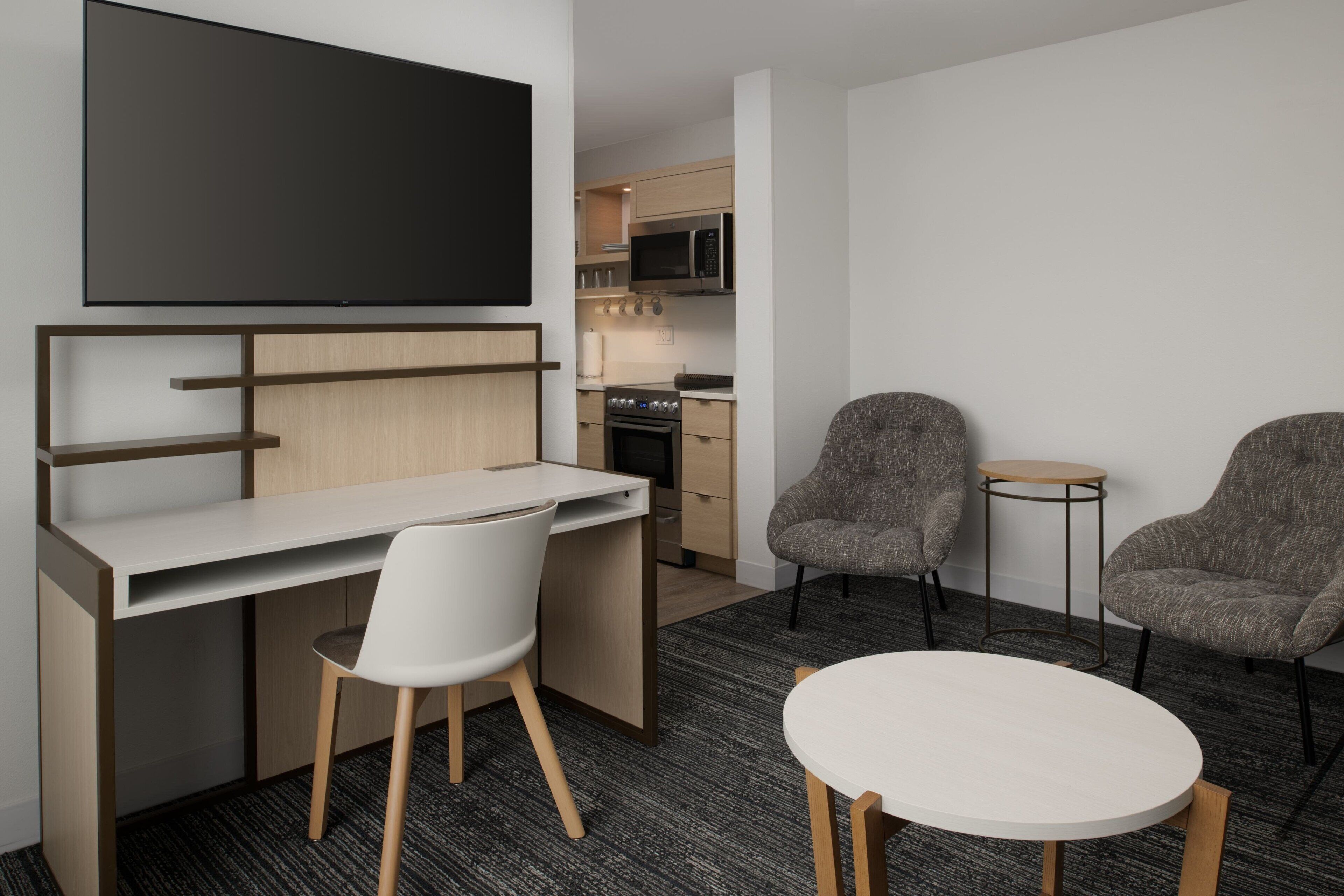 A modern hotel room with a flat-screen TV mounted above a desk, two armchairs, a small round table, and a kitchenette in the background.
