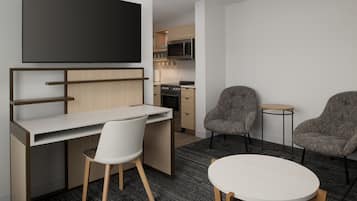 A modern hotel room with a flat-screen TV mounted above a desk, two armchairs, a small round table, and a kitchenette in the background.