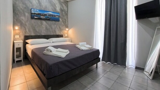 A neatly made bed with white linens and towels, a small bedside table, a wall-mounted TV, and a painting above the bed. - GIORGIA'S HOUSE - VACATION HOME (Catania)
