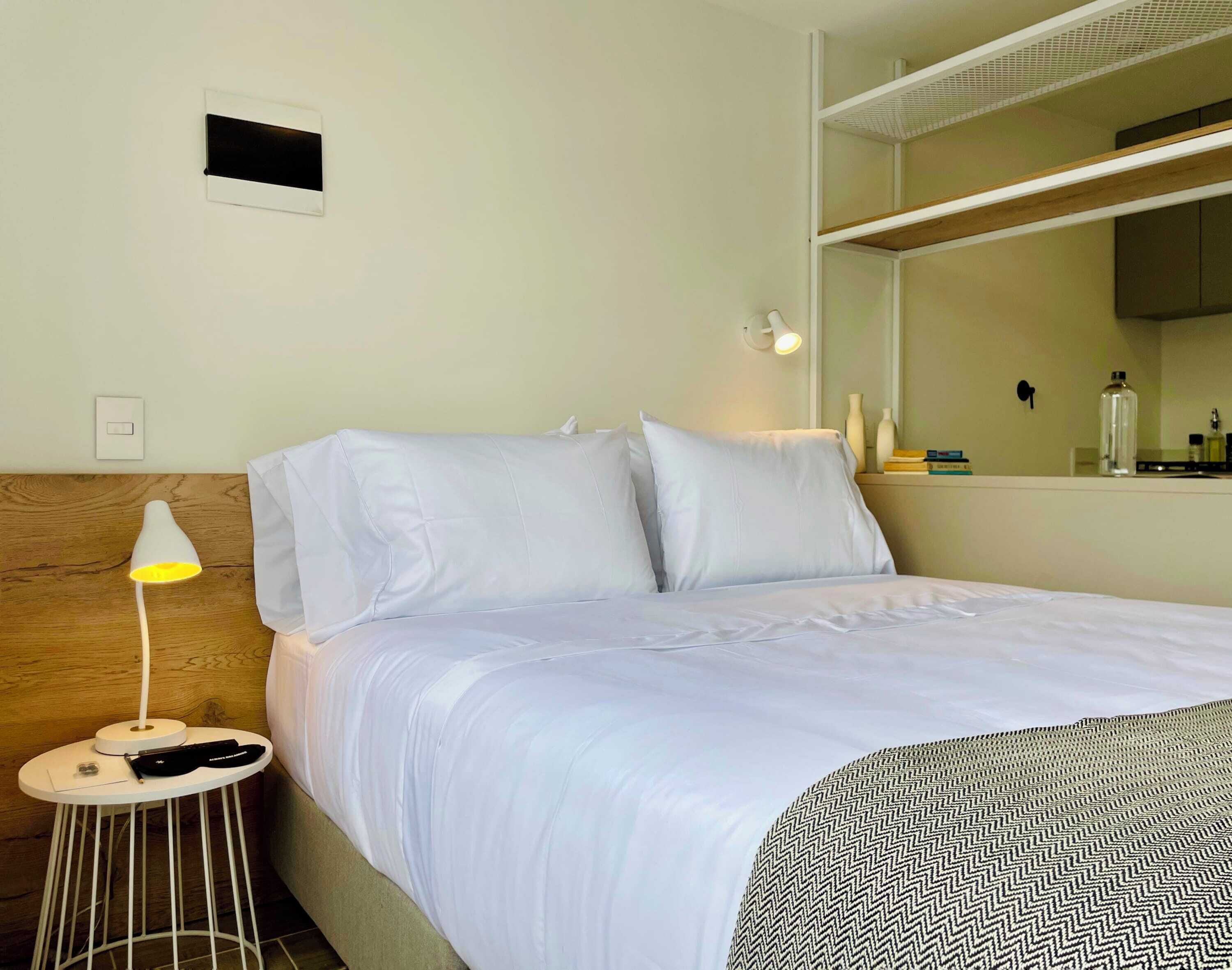 Een hotelkamer met een bed, een nachtkastje met een lamp, een spiegel en een klein plankje met toiletartikelen.