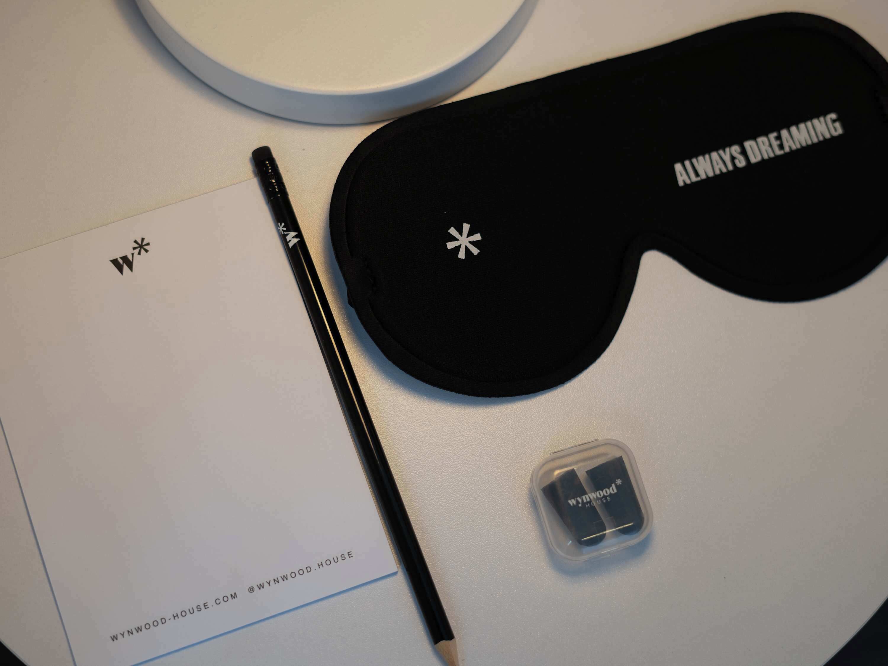 Eine schwarze Augenmaske mit dem Aufdruck "ALWAYS DREAMING", ein schwarzer Stift, ein weißes Notizbuch mit "WYNNWOOD-HOUSE.COM @WYNNWOOD.HOUSE" und eine kleine Schachtel mit "WYNNWOOD HOUSE".