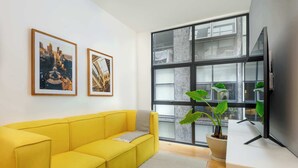 Um sofá amarelo vibrante, uma janela ampla com vista e uma TV de tela plana fixada na parede.