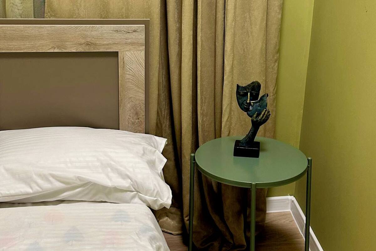 A bedroom with a bed, a green side table with a sculpture, and a window with curtains.