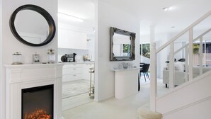 Un intérieur moderne et épuré, aux couleurs claires, avec une cheminée, un miroir rond et un coin repas offrant une vue sur l’extérieur.