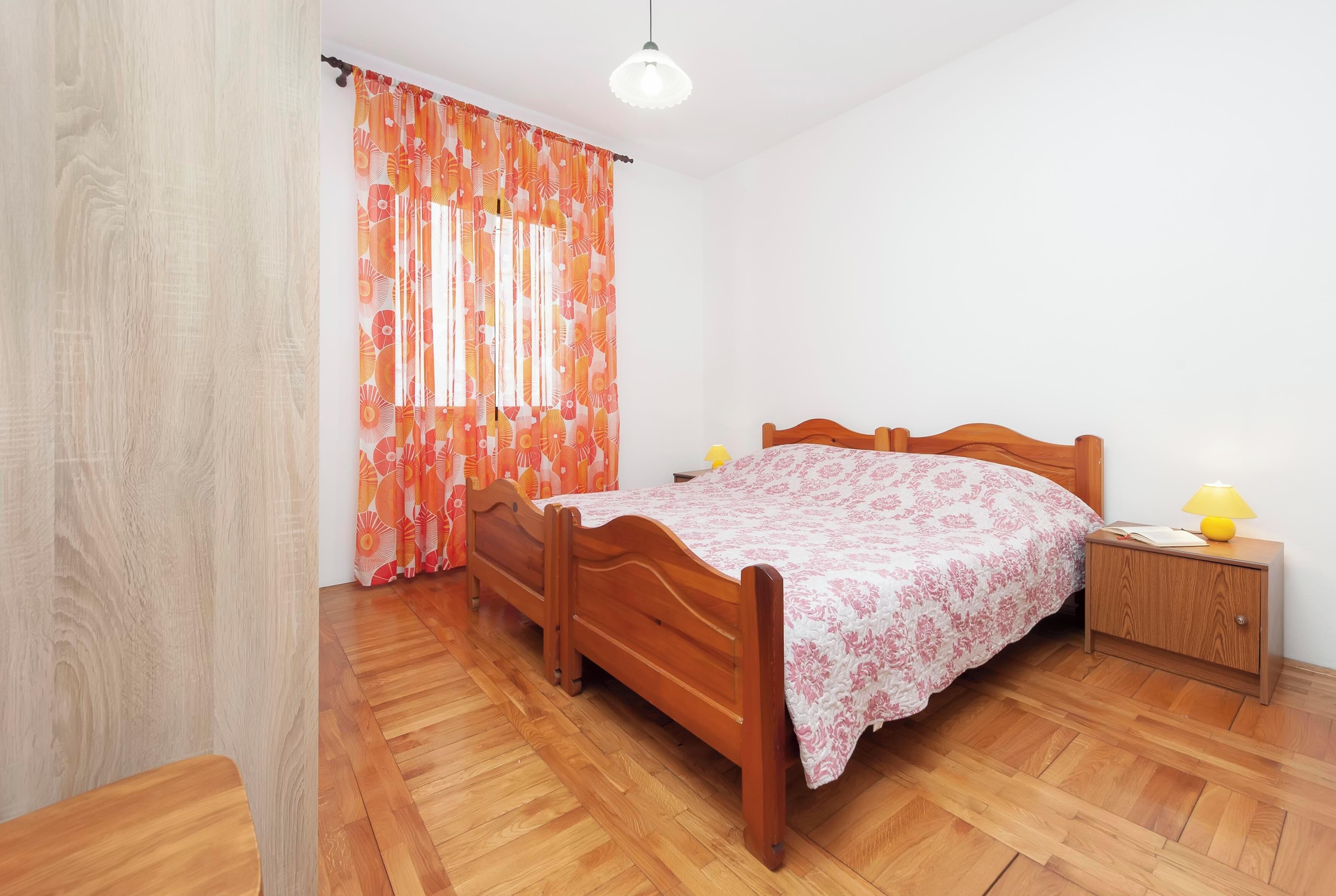 Ein Schlafzimmer mit einem hölzernen Bett, einem Nachttisch, einer Lampe und einem Fenster mit orangefarbenen Vorhängen.