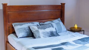 A wooden bed with a headboard, two patterned pillows, a white bedspread, and a small bedside table with a candle. - Claris Cozy Homes Near US Mall (Kampala)
