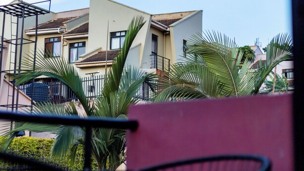 A view of a residential building with a balcony, palm trees, and a red wall. - Claris Cozy Homes Near US Mall (Kampala)